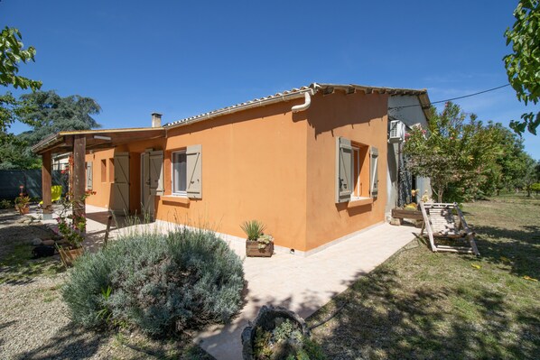 Une maison à deux étages, avec un toit en tuiles, un porche à l’avant et un petit jardin où poussent différentes espèces de plantes.