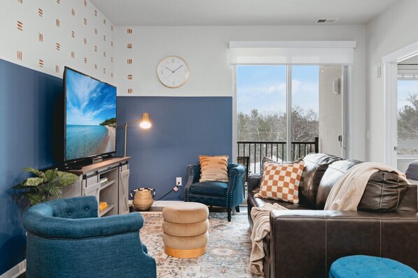 A modern living room with a large flat-screen TV, a blue armchair, a brown leather sofa, and a patterned rug. - *NEW* Great Escape 404 2BR Condo Near Downtown and Bay (Traverse City)