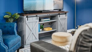Un salón con un televisor de pantalla plana grande sobre un soporte de madera, un sillón azul, un reposapiés redondo pequeño y una lámpara de pie.
