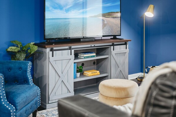 A living room with a large flat-screen TV on a wooden stand, a blue armchair, a small round ottoman, and a floor lamp. - *NEW* Great Escape 404 2BR Condo Near Downtown and Bay (Traverse City)