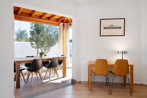 A dining area with a wooden table and chairs, a framed picture on the wall, and a view of trees outside.