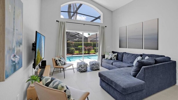 A spacious living room with a large grey sofa, a coffee table, and a TV mounted on the wall. There is a window with a view of a swimming pool and greenery outside. - Super Spacious House w/ Private Pool and Game Room (Kissimmee)
