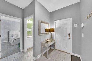 Un passadís amb una porta blanca, un mirall, un tocador amb una làmpada i un llit a l'habitació veïna.