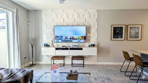 A modern living room with a flat-screen TV mounted on a white wall with geometric patterns. There are two brown leather ottomans, a glass coffee table with a book, and a framed sign on the floor. - Modern 4BR Townhome with Private Pool for 10 (Kissimmee)