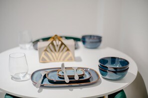 Una mesa redonda con superficie blanca, un vaso transparente, un juego de platos azules y marrones, un par de cubiertos blancos y un cuenco azul.