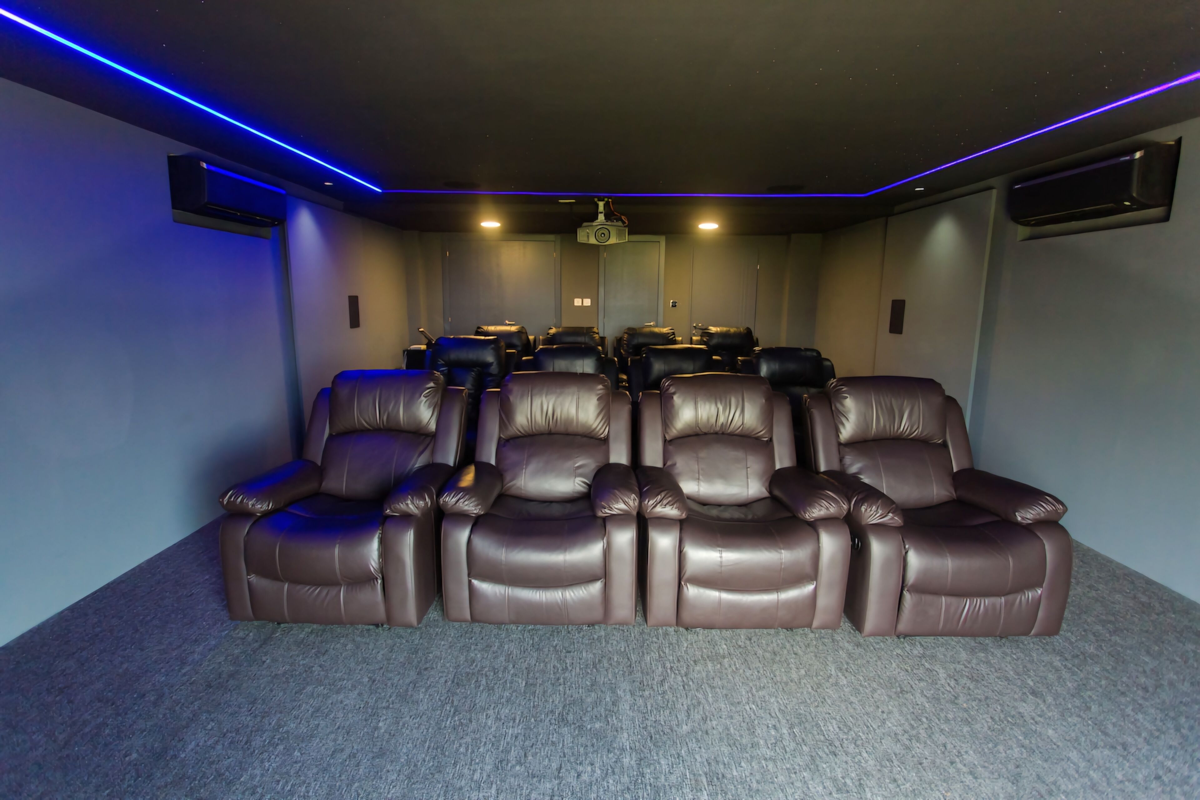 Ein Heimkinozimmer mit einer Reihe brauner Ledersessel, einem Deckenprojektor und blauer LED-Beleuchtung.