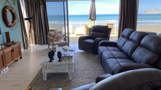 A living room with a view of the ocean, a sofa, a chair, a coffee table, and a wooden cabinet. - Ultimate Beachfront - Family Friendly with games room and multiple decks (Waimarama)