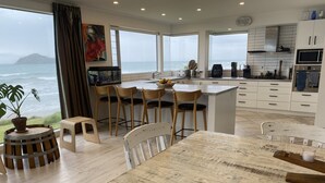 A modern kitchen with a large island, wooden chairs, and a view of the ocean. - Ultimate Beachfront - Family Friendly with games room and multiple decks (Waimarama)