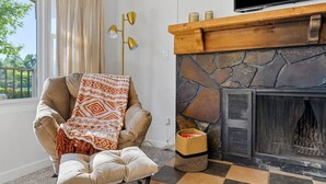 A cozy living room with a stone fireplace, a comfortable armchair with a patterned throw, and a view of greenery outside. - Home located close to Downtown (Spearfish)