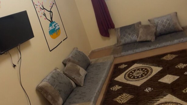 A living room with a sofa, a TV mounted on the wall, a patterned rug, and a ceiling light fixture. - Résidence Mouna (Nouadhibou)