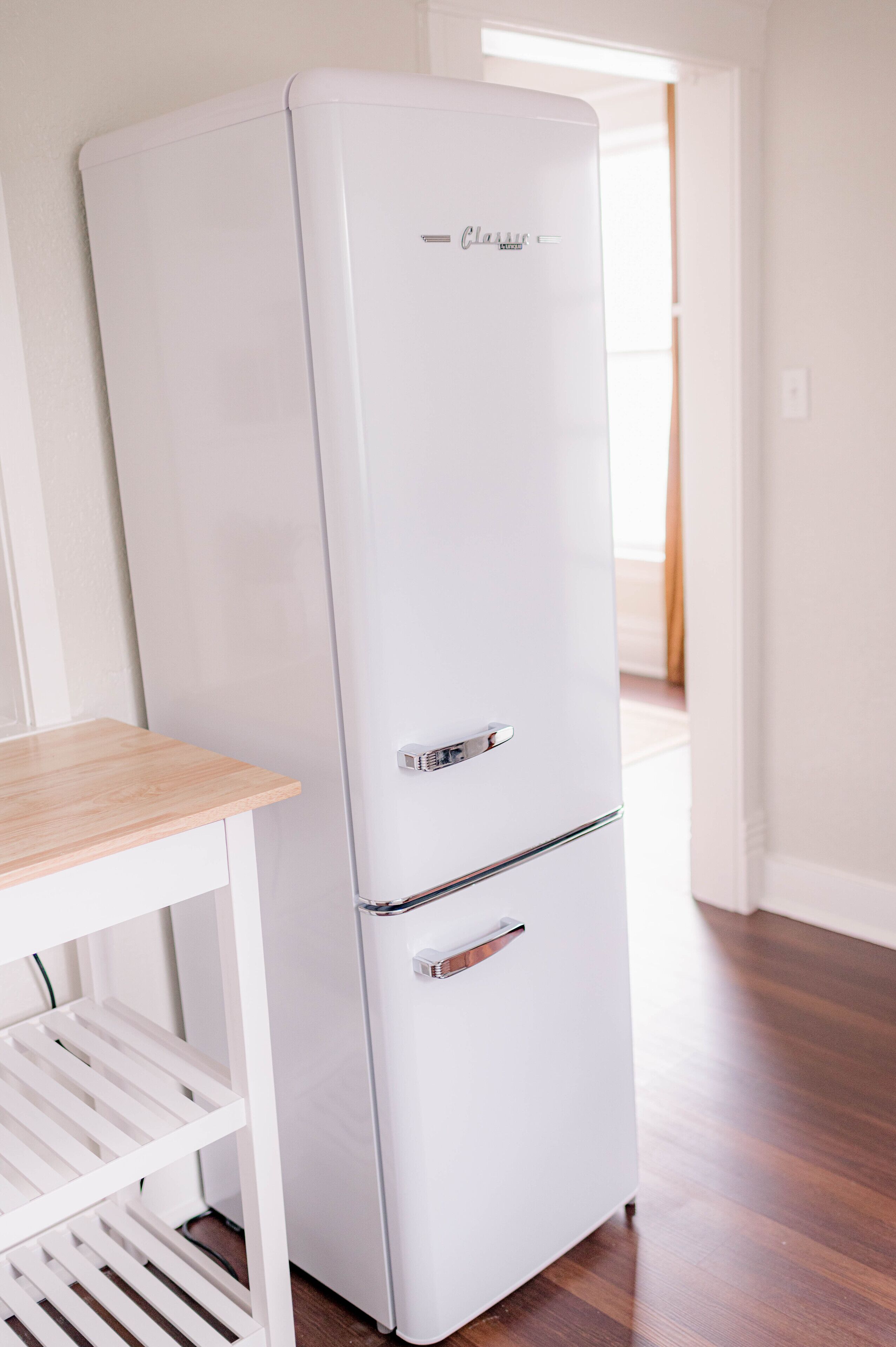 A white Classic refrigerator in a room with wooden flooring and a white table with shelves.