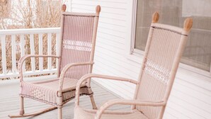 Two wicker rocking chairs on a wooden porch with a white exterior wall and windows.
