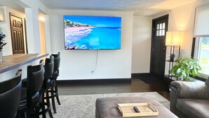 A living room with a flat-screen TV mounted on the wall, a bar area with chairs, a grey sofa, and a wooden tray with a remote control. - Hot Tub & Cloud Ceiling House (Montville)