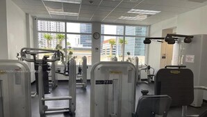Une salle de sport équipée de divers appareils d’exercice, de grandes fenêtres offrant une vue sur la ville et un panneau indiquant une zone de service à température contrôlée.