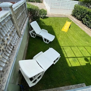 A small, enclosed backyard with artificial grass, two white lounge chairs, and a yellow slide.