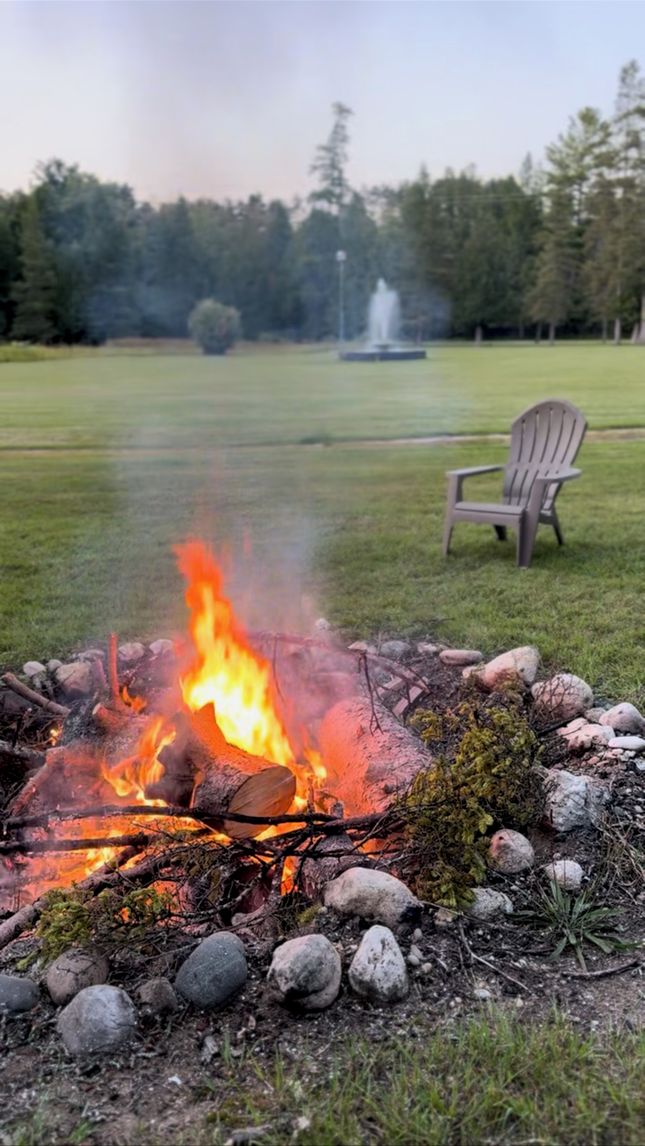 Un feu de camp entouré de rochers et de bûches, une chaise et une fontaine, le tout situé dans un champ herbeux.