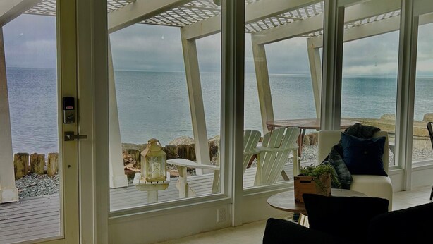 A room with large windows overlooking a beach. There is a door leading outside, a small table with chairs, and a potted plant. - Modern Mid-Century Cabin, on the beach, with views of the San Juan Islands! (Oak Harbor)