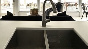 A modern kitchen with a large sink, a hanging light fixture, and large windows offering a view of the outdoors. - Modern Mid-Century Cabin, on the beach, with views of the San Juan Islands! (Oak Harbor)