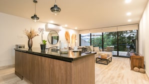 A modern kitchen with a wooden island, a dining area with a sofa and table, and large windows offering a view of the outdoors.