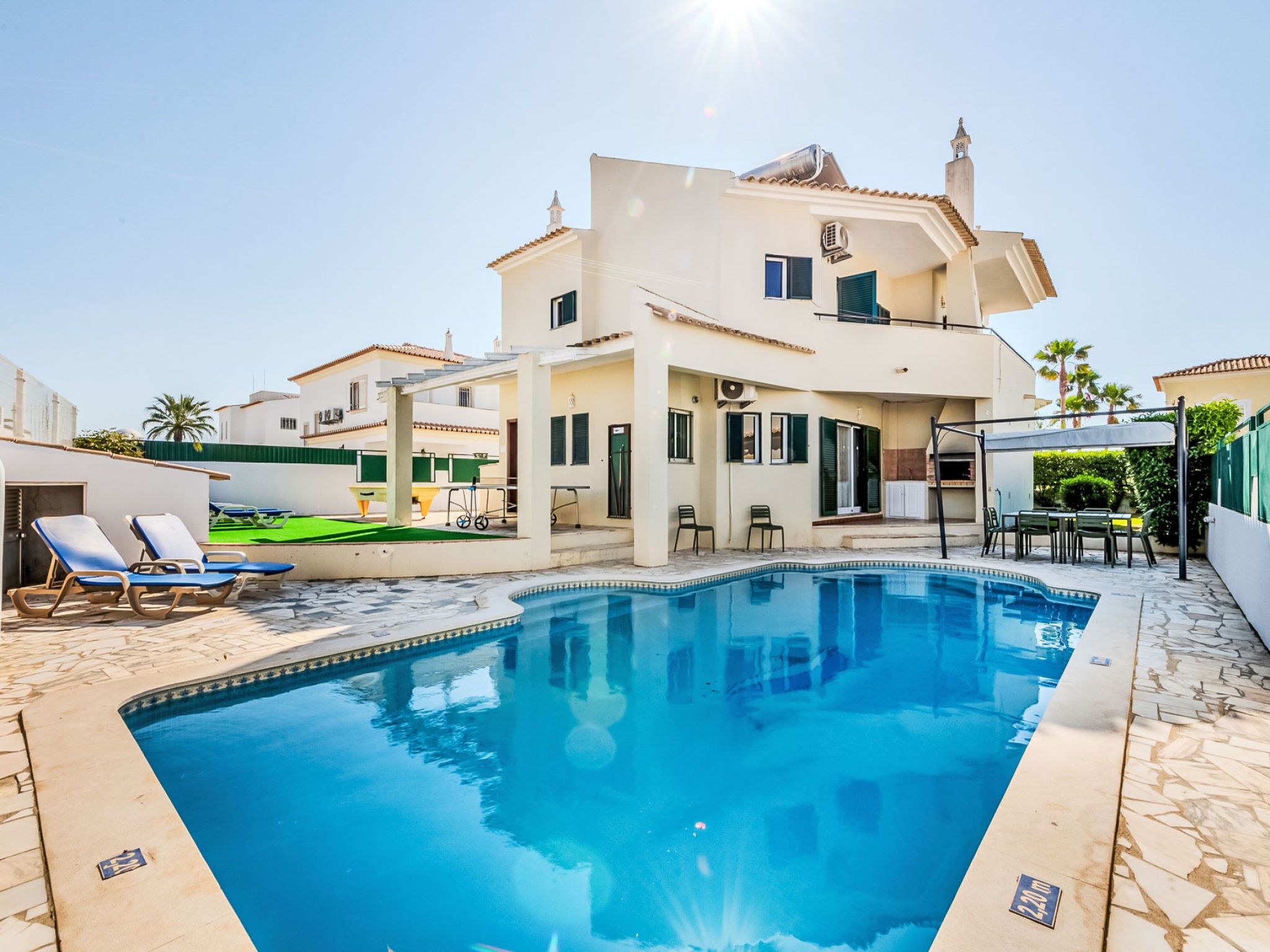 Une piscine avec des chaises longues, un espace salon couvert et un bâtiment à deux étages doté d’un balcon.