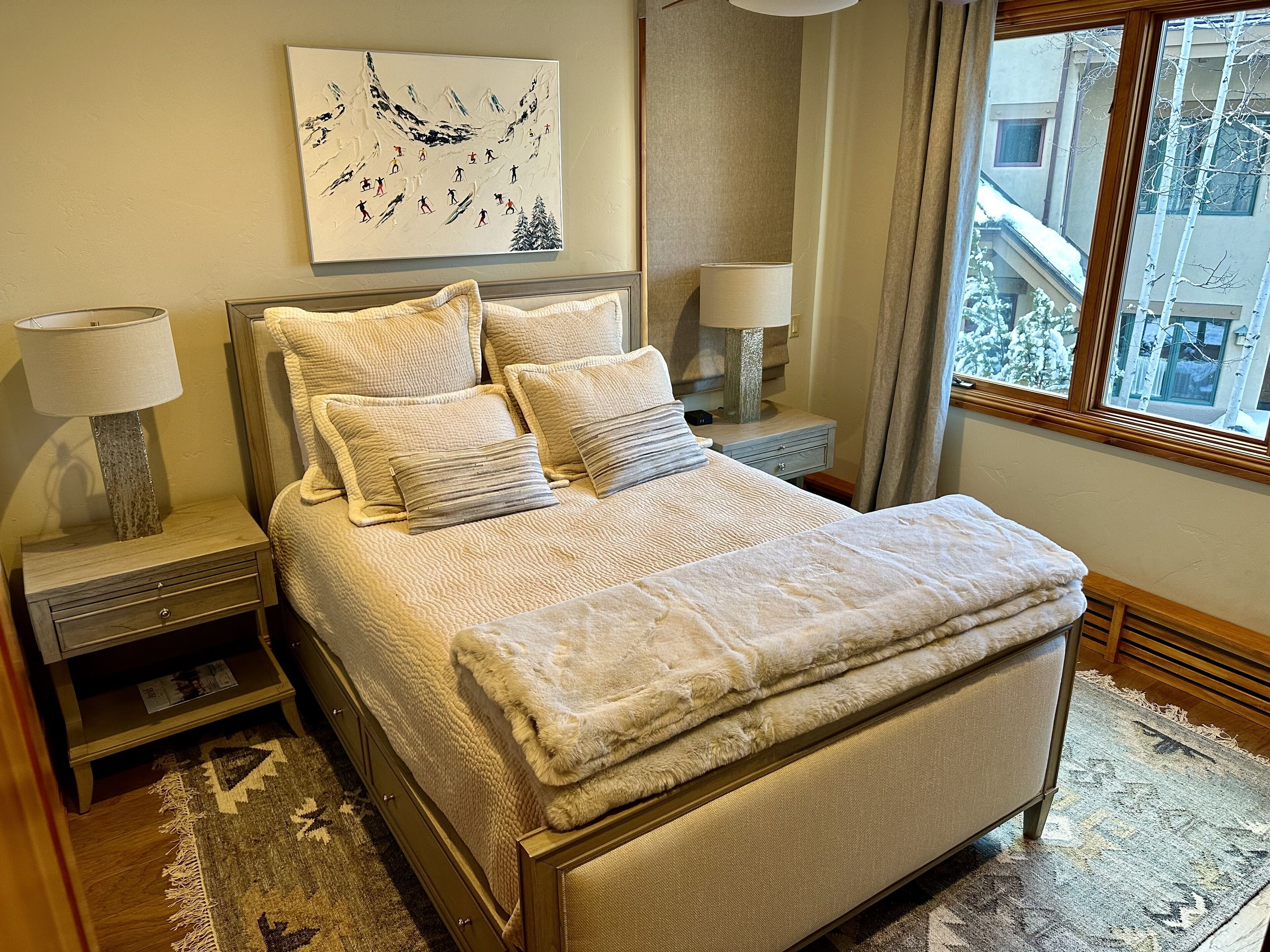 A bedroom with a large bed, two bedside tables with lamps, a wall-mounted painting, and a view of a snowy building through a window.