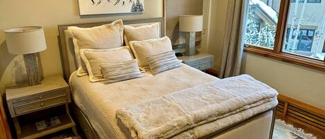 A bedroom with a large bed, two bedside tables with lamps, a wall-mounted painting, and a view of a snowy building through a window.