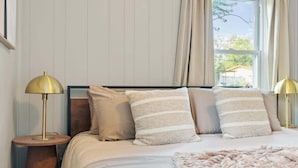 Ein Schlafzimmer mit einem Bett, zwei Nachttischlampen, einem Fenster mit Vorhängen und Blick ins Freie.