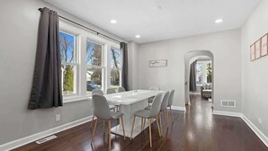 A dining area with a white table and grey chairs, a window with curtains, and a hallway leading to another room. - Bungalow w/ Game Room | LCP Collection (Morton Grove)