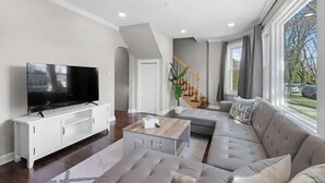 A modern living room with a flat-screen TV, a white entertainment unit, a grey sofa, a coffee table, and a staircase leading to an upper level. - Bungalow w/ Game Room | LCP Collection (Morton Grove)