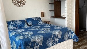 A bedroom with a white brick headboard, a blue floral bedspread, a wall-mounted air conditioner, a decorative wall piece, and a wooden shelf with a mirror. - Altos da Lagoa-praia do Rosa, Ibiraquera -studio 2 (imbituba)