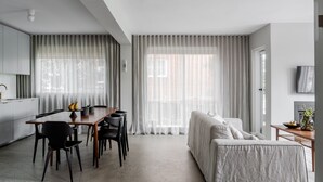 Ein modernes, minimalistisches Wohnzimmer mit Essbereich, großen Fenstern mit transparenten Vorhängen, einer Couch und einem flachbildfernseher an der Wand.