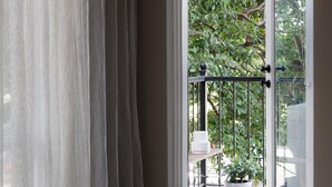 Blick durch ein Fenster mit durchscheinenden Vorhängen, einem Metallgeländer, einem Topfpflanze und einem weißen Tisch.