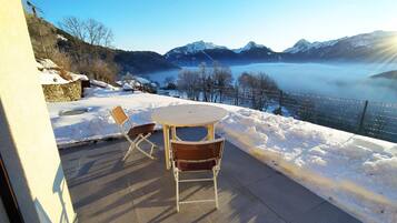 A patio with a table and chairs, overlooking a snowy landscape with mountains and a lake.
