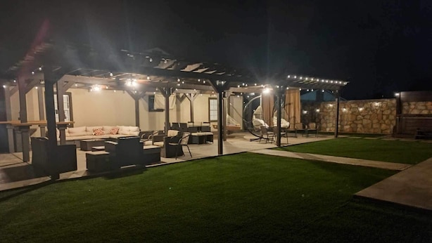 Un espace extérieur aménagé en patio, doté d’une pergola, de sièges et d’une balançoire, le tout entouré d’un mur de pierres et bordé d’une pelouse bien entretenue.