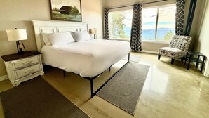 Um quarto com cama, mesa de cabeceira, cadeira, janela com cortinas e um quadro na parede.