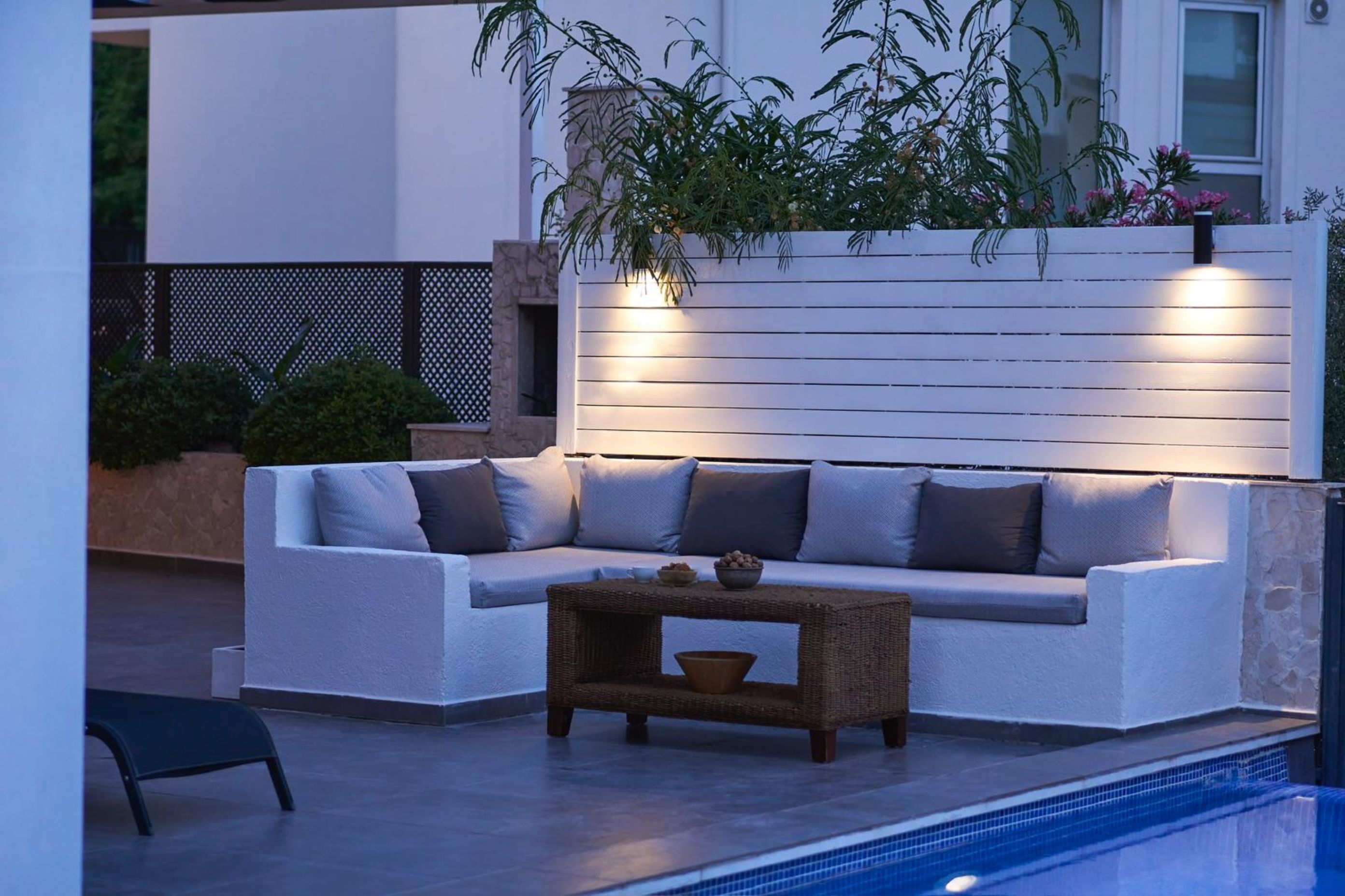 A modern outdoor seating area with a white sofa, a wicker coffee table, and a pool.