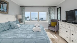 A bedroom with a large bed, a TV, a chair, and a window with curtains.