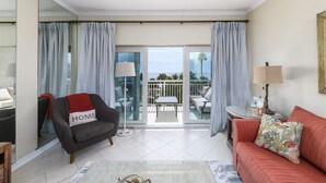 A living room with a sofa, armchair, and a small table with a lamp. There is a balcony with a view of the sea.