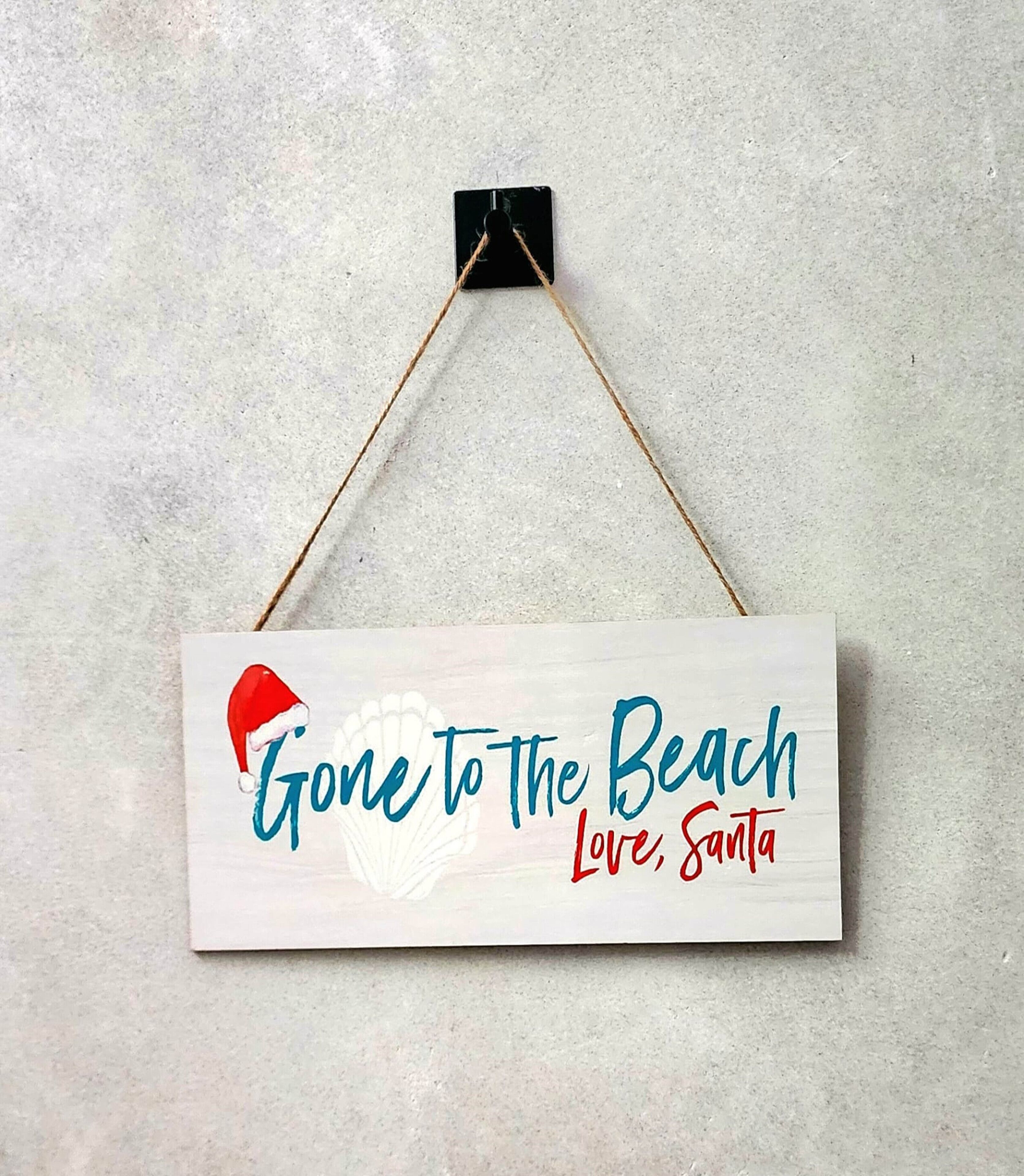 Een rechthoekig bord met een kerstmuts en de tekst 'Gone to the Beach Love, Santa' hangt aan een muur.