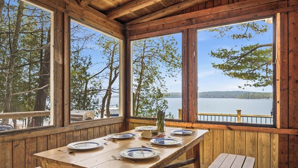 A wooden cabin with a dining table set for four, large windows overlooking a lake, and a wooden deck.