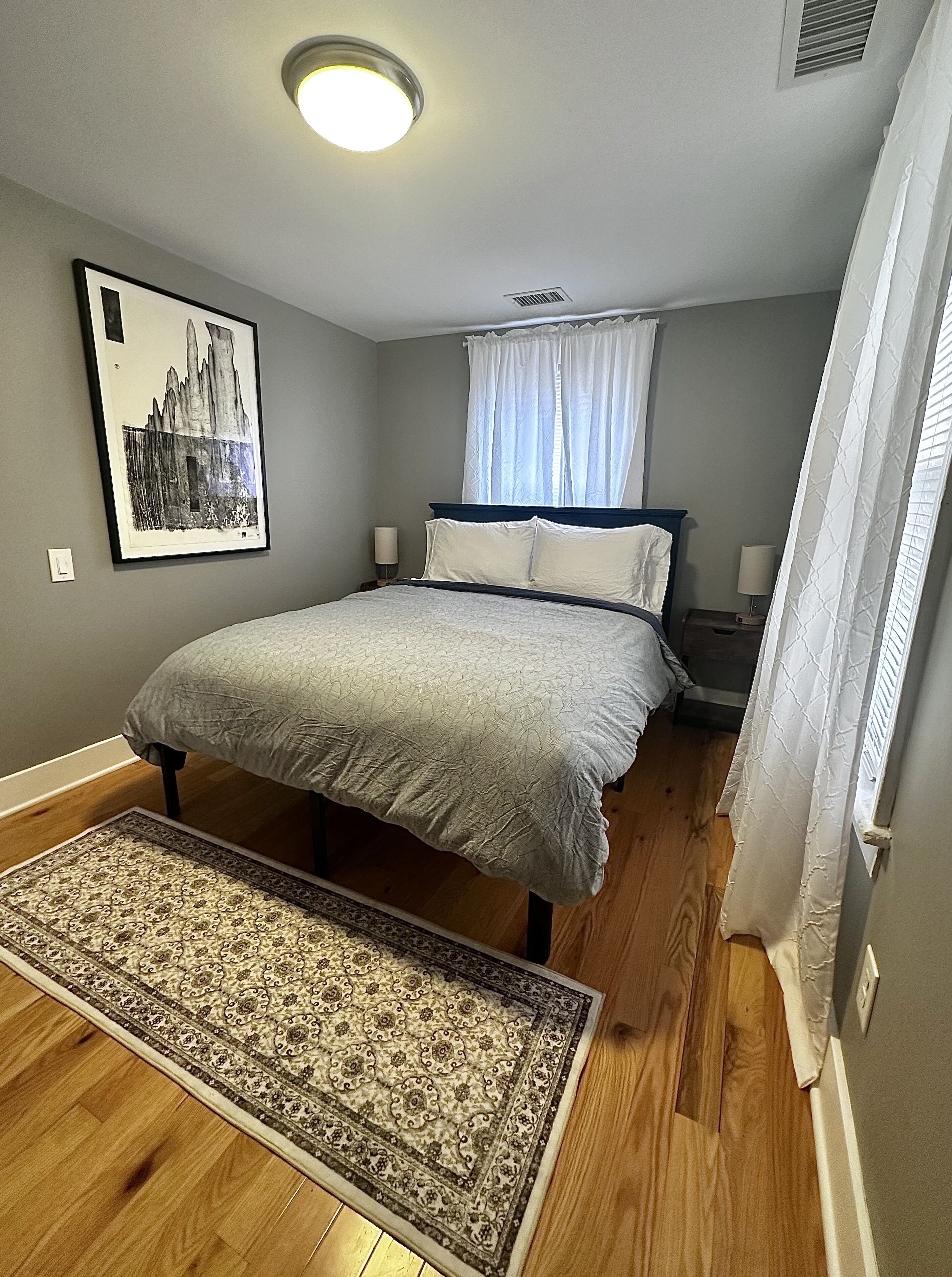 Un dormitorio con cama, alfombra, un cuadro en la pared y una ventana con cortinas.