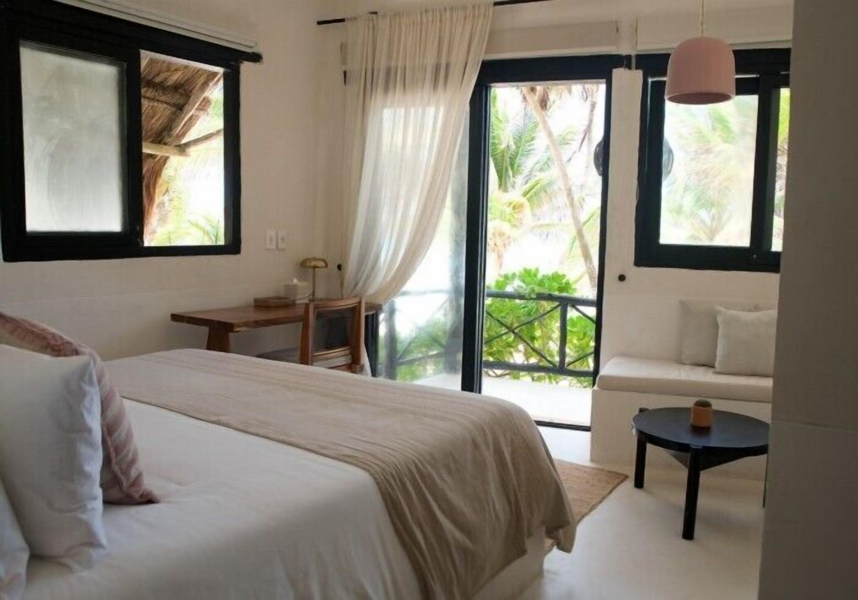 Un dormitorio con cama, escritorio, silla, una mesita y un balcón con vistas a la vegetación.