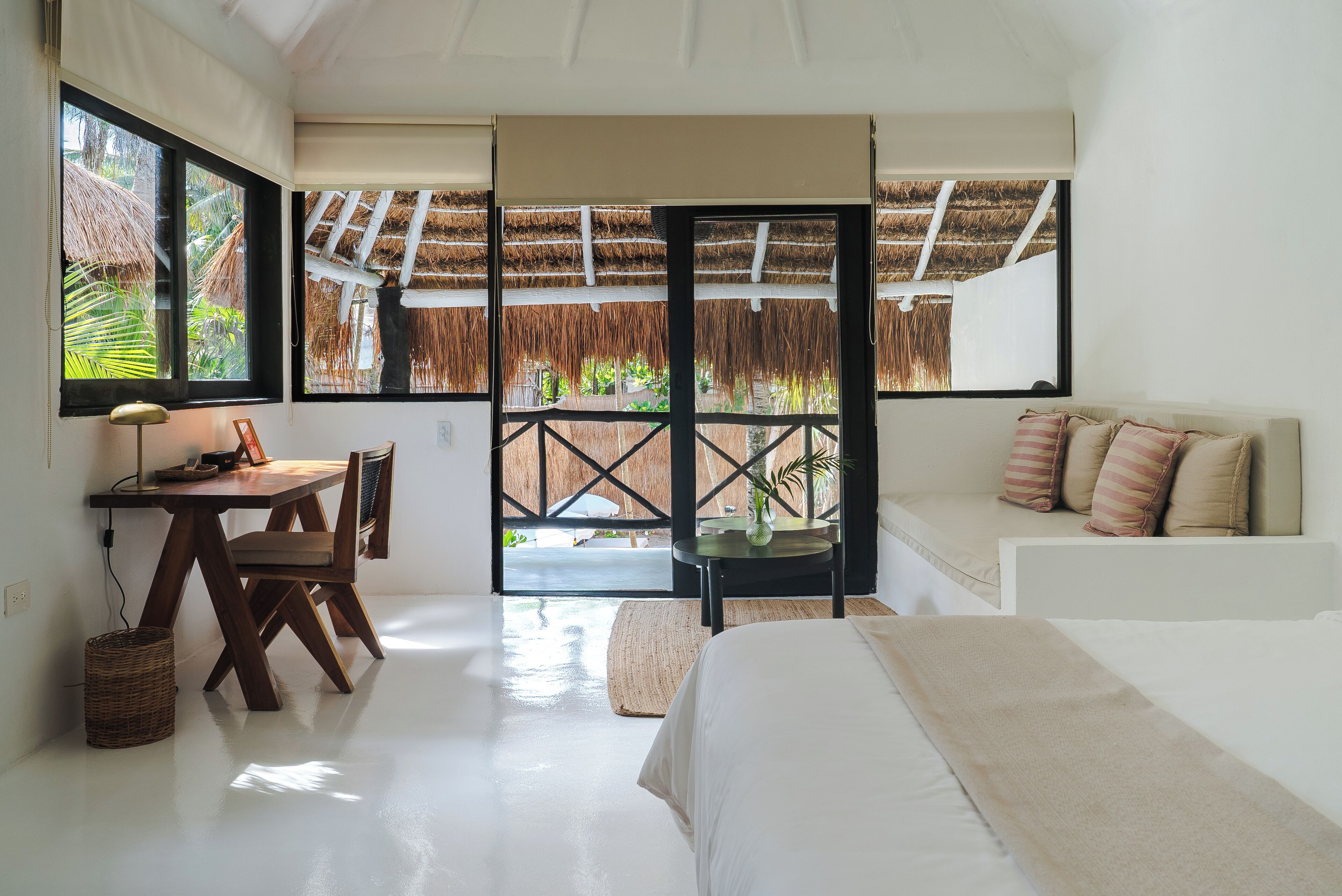 A bedroom with a bed, a desk, and a chair. There is a window with a view of a thatched roof and greenery.