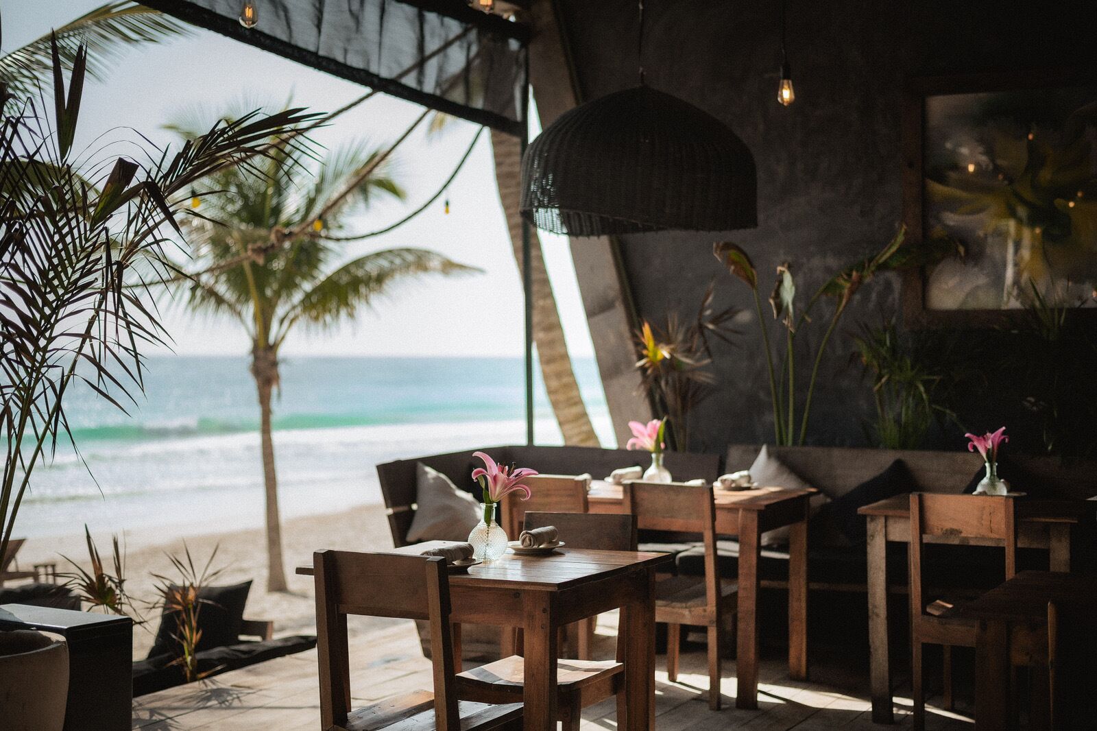 A seaside restaurant with wooden furniture, a view of the ocean, and tropical plants.