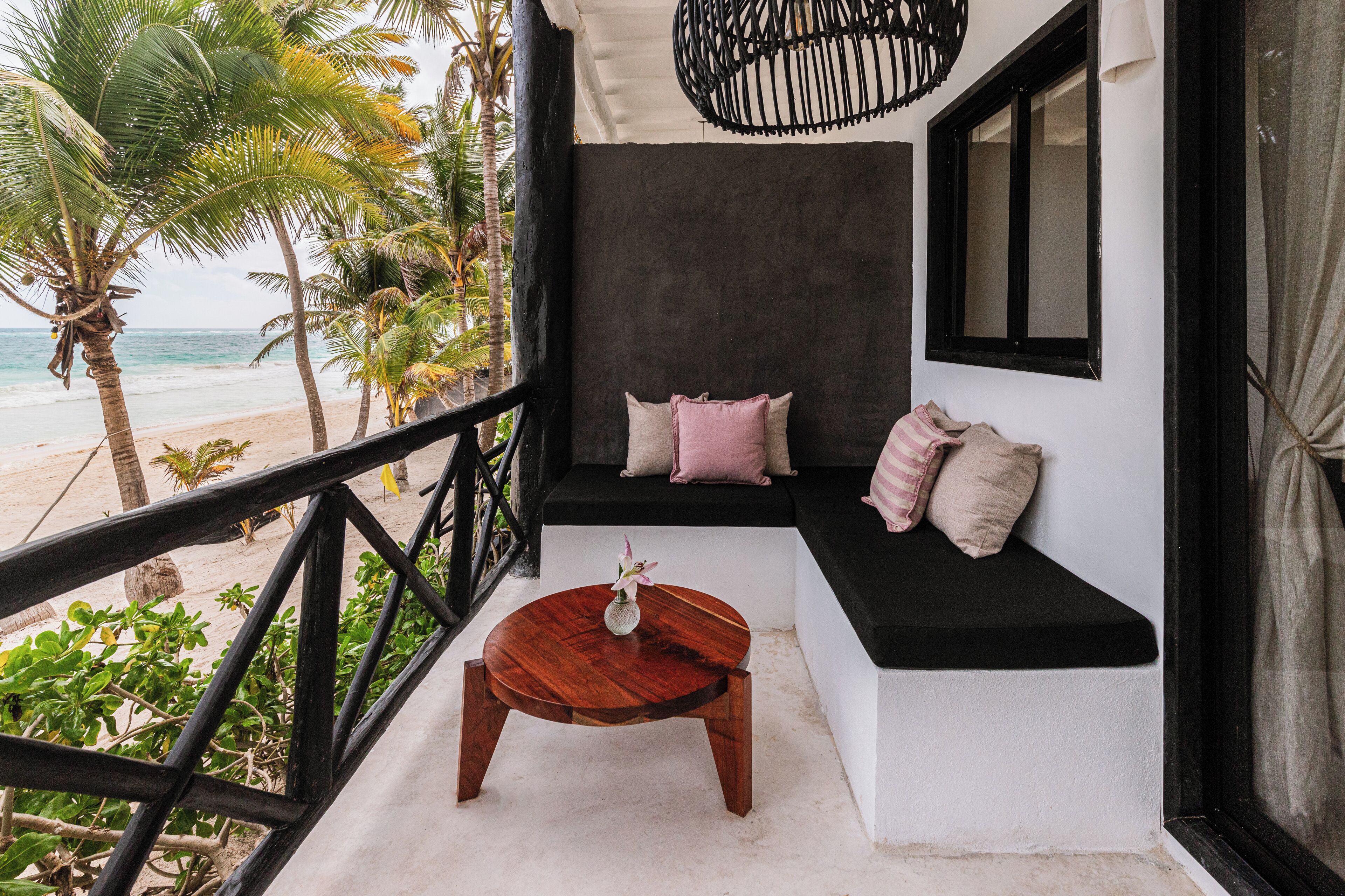 Un balcone con un tavolo di legno, una panca imbottita con cuscini e vista sulla spiaggia e sulle palme.