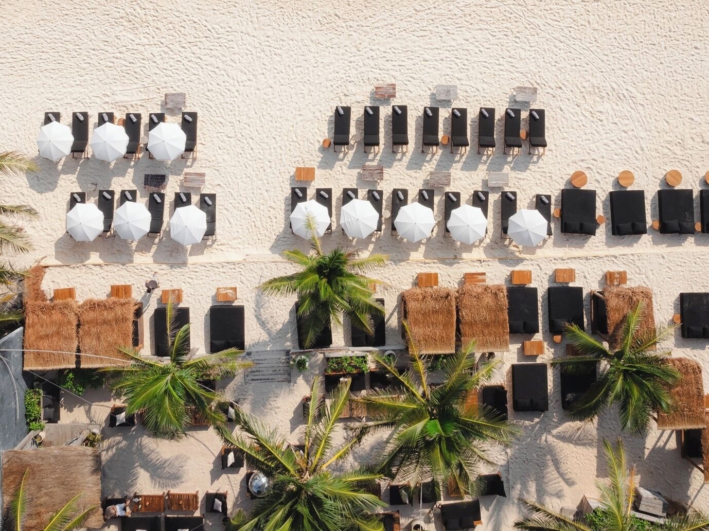 A beachside lounge with rows of sun loungers and umbrellas, surrounded by palm trees and thatched structures.