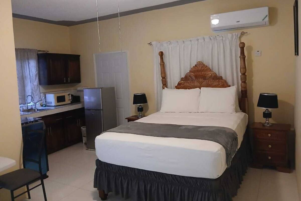 A hotel room with a bed, a desk, a microwave, a refrigerator, and an air conditioning unit.