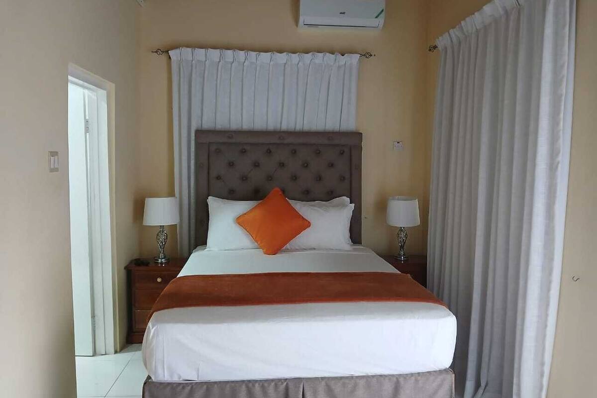 A hotel room with a large bed, two bedside tables with lamps, a window with curtains, and an air conditioning unit on the wall.
