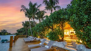 A wooden deck with lounge chairs, a pool, and a building with warm lighting. - Heated pool / Lush Tropical Surrounding / Castaway Key / RESlDENCES (Fort Lauderdale)