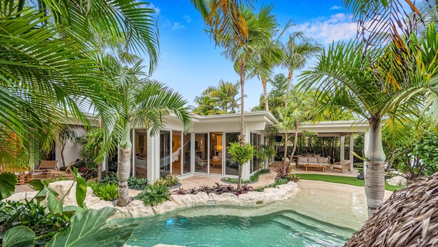 A pool surrounded by tropical plants and a modern house with a covered patio. - Heated pool / Lush Tropical Surrounding / Castaway Key / RESlDENCES (Fort Lauderdale)
